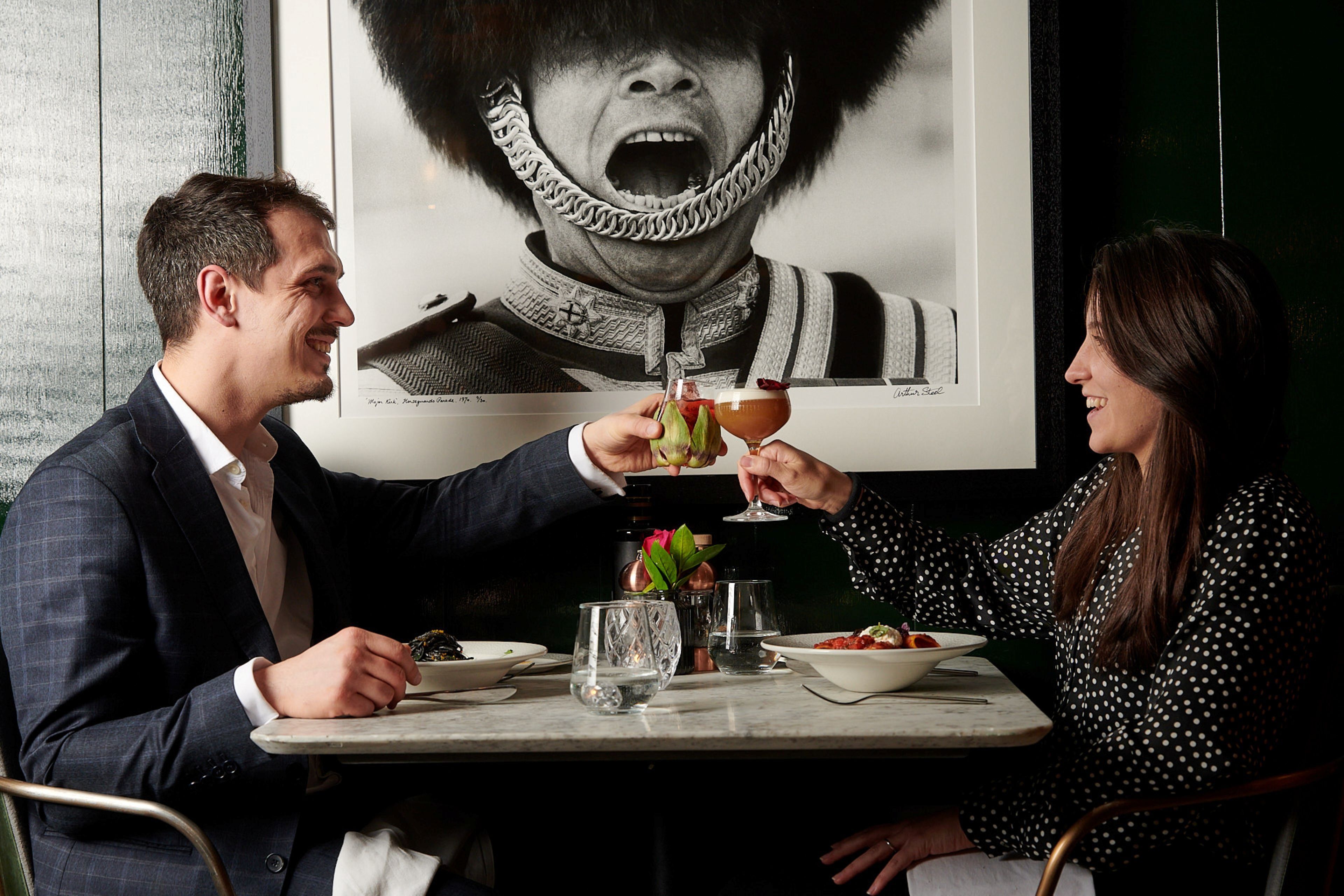 A couple dining at a table with a drink in each hand, a plate of food, and a vase with flowers.