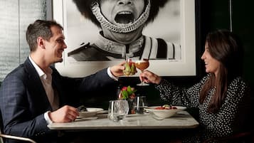 A couple dining at a table with a drink in each hand, a plate of food, and a vase with flowers.
