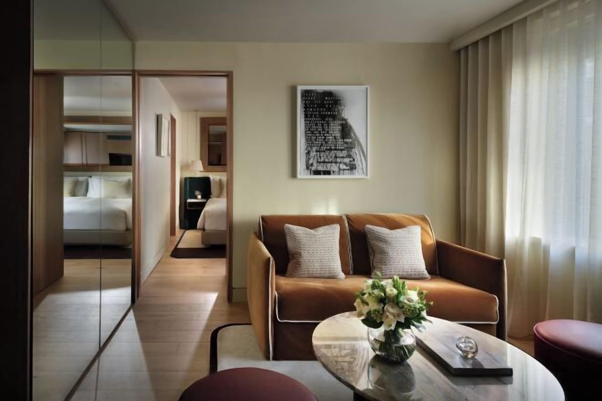 A hotel room with a sofa, a coffee table with flowers, a framed picture on the wall, and a view of another room through a glass door.