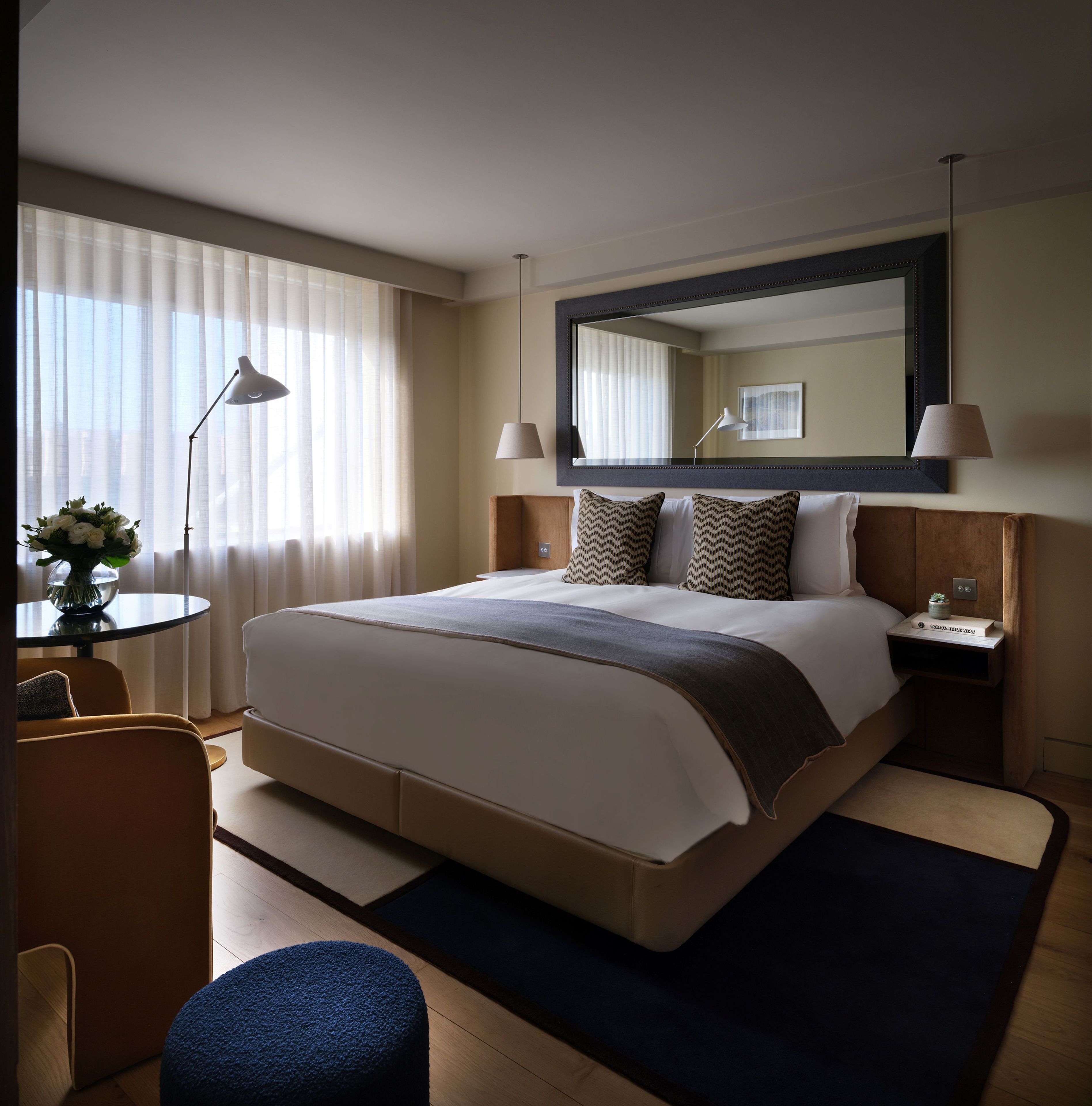 A hotel room with a large bed, bedside lamps, a round table with a vase of flowers, and a chair.