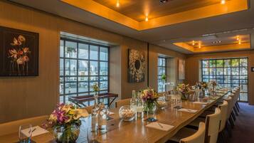A long conference table with chairs, glassware, and floral centerpieces in a well-lit room with large windows and artwork on the walls.