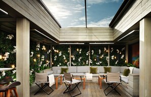Un área de estar al aire libre con diseño moderno, que cuenta con un techo de madera y una pared decorada con luces colgantes y plantas.
