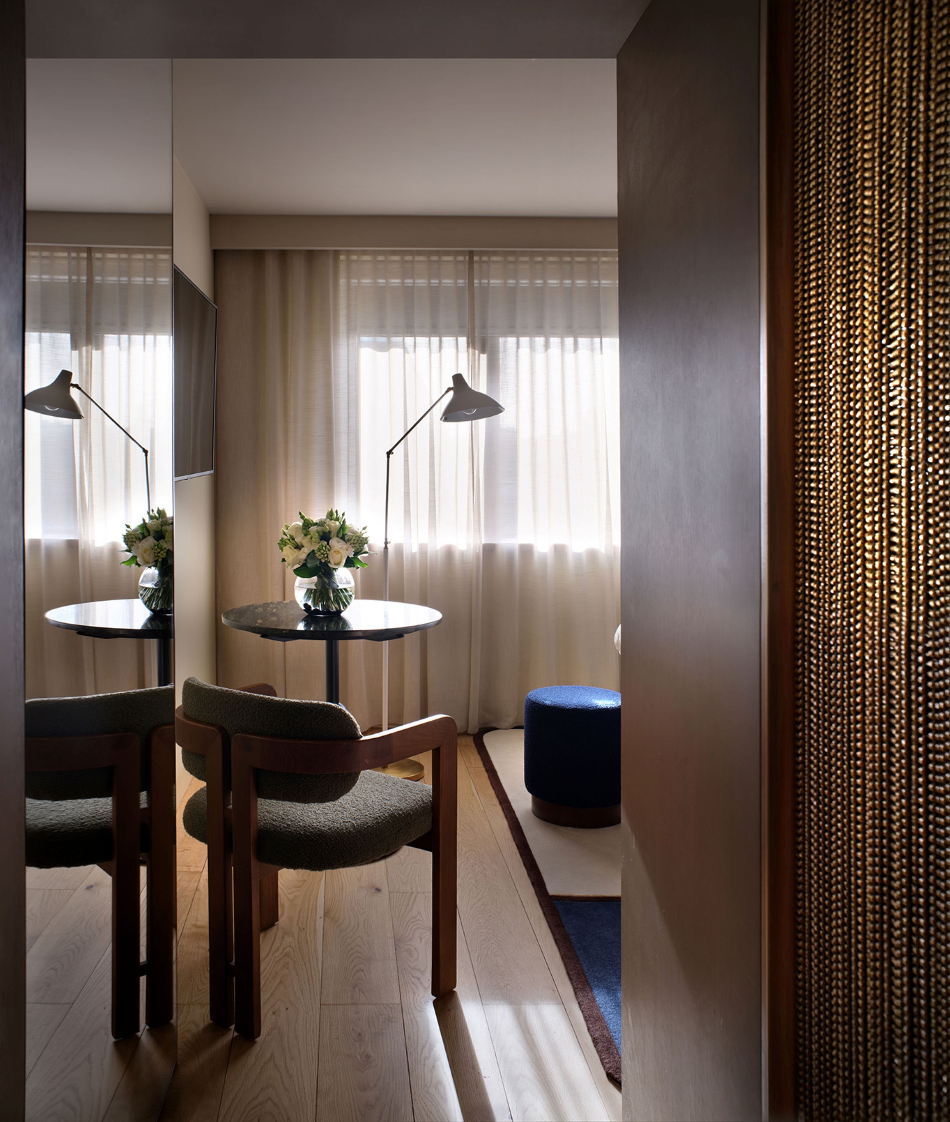 Quarto de hotel com uma mesa redonda, duas cadeiras, um banquinho azul pequeno, um vaso com flores e um espelho grande na parede.