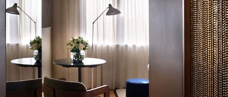 A hotel room with a round table, two chairs, a small blue ottoman, a vase with flowers, and a large mirror on the wall.