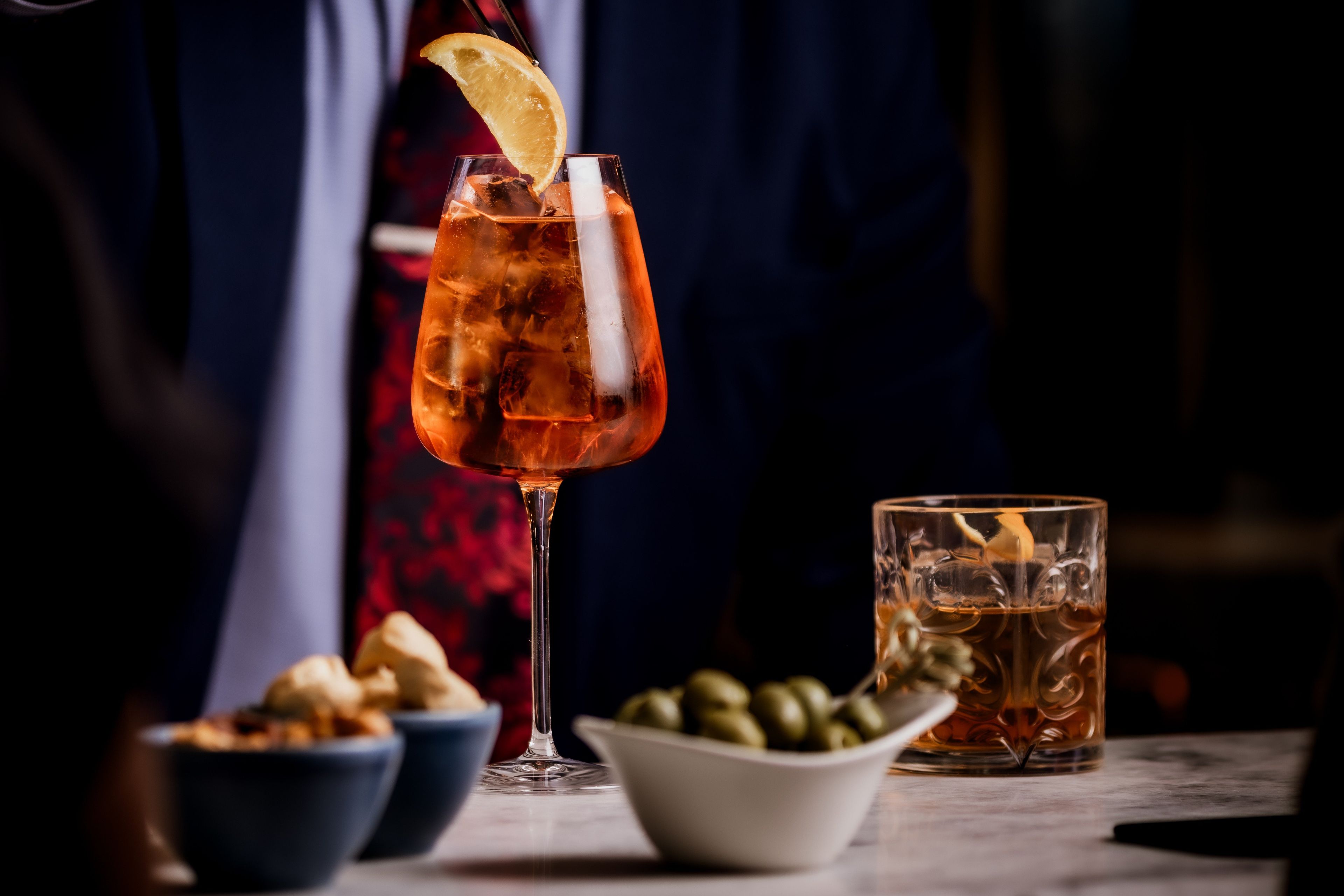 Una copa de cóctel con rodaja de limón, cubitos de hielo y una bebida oscura, acompañada de un vaso de té helado y recipientes con aceitunas y frutos secos.