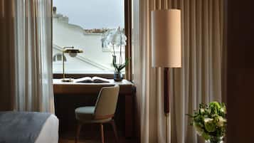 A hotel room with a bed, a desk with a chair, a standing lamp, and a view of the cityscape.