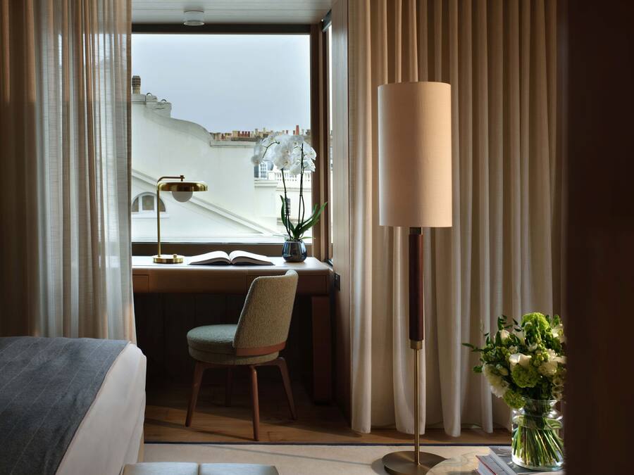 A hotel room with a bed, a desk with a chair, a standing lamp, and a view of the cityscape.
