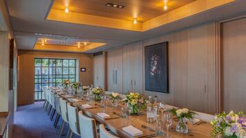 A long conference table set with chairs, floral centerpieces, and glassware, in a room with wooden paneling and a large window.
