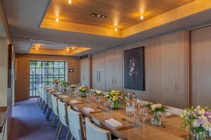 Una larga mesa de conferencias dispuesta con sillas, centros de mesa florales y cristalería, en una sala con paneles de madera y un ventanal grande.