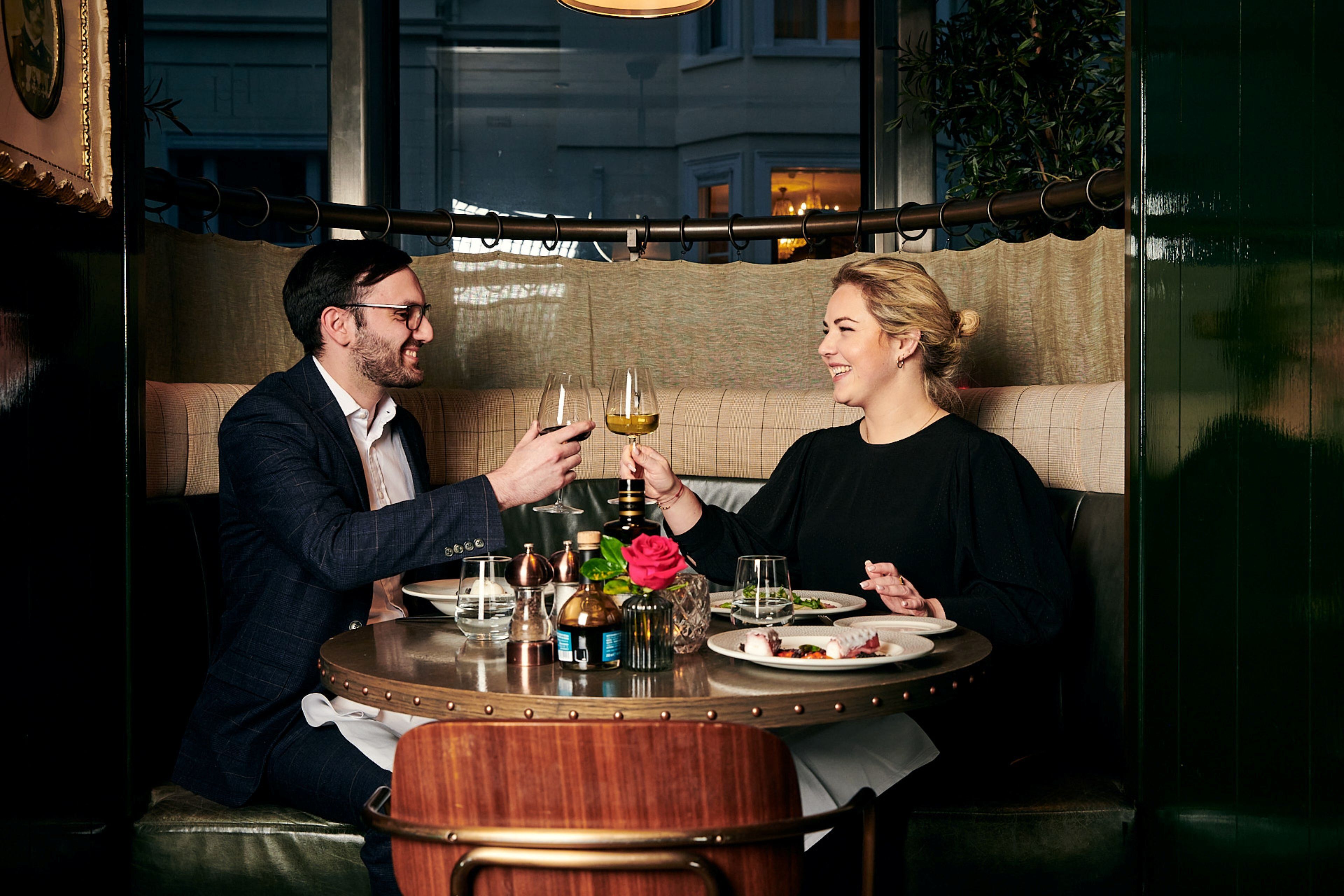 Um casal sentado à mesa redonda com uma garrafa de vinho, um copo e uma rosa.