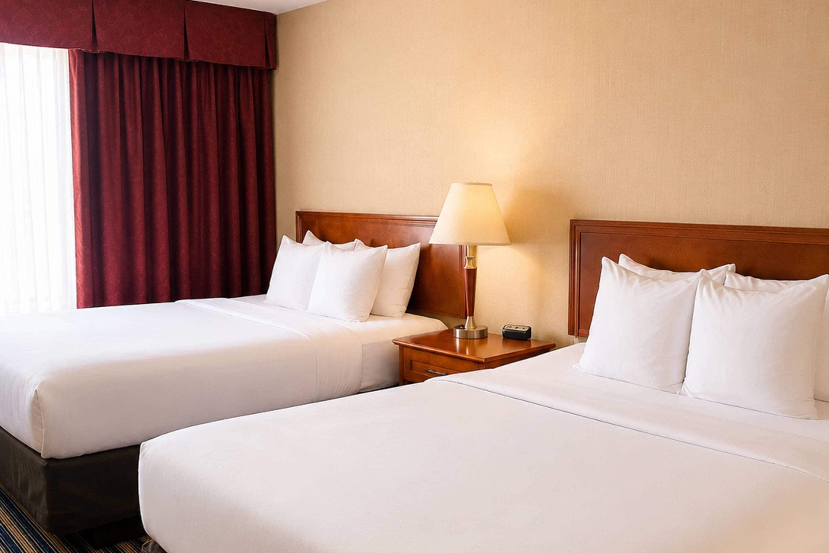 A hotel room with two beds, a nightstand with a lamp, and a window with red curtains.
