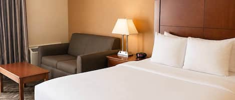 A hotel room with a large bed, a chair, a small table, a nightstand with a lamp, and a clock.