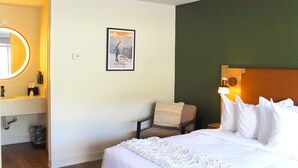 A hotel room with a bed, a chair, a nightstand, a mirror, a framed picture, and a wall-mounted light.