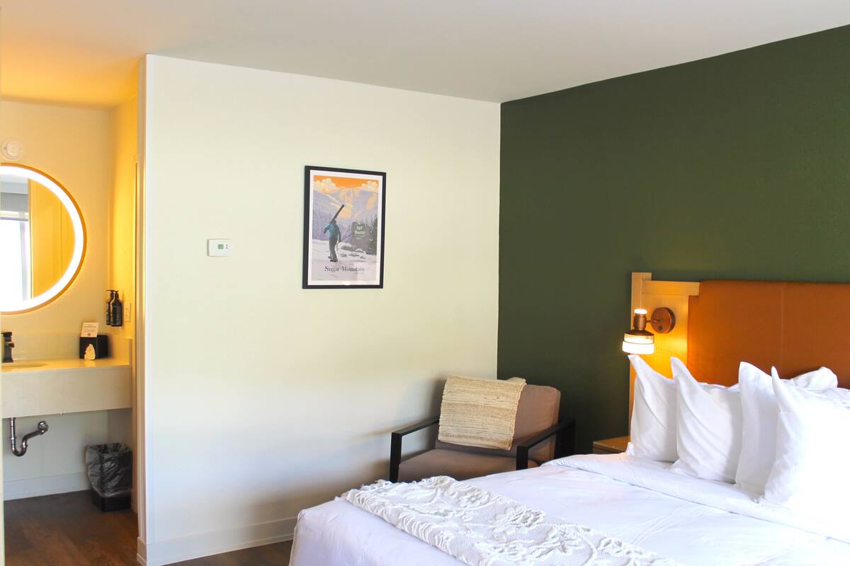 A hotel room with a bed, a chair, a nightstand, a mirror, a framed picture, and a wall-mounted light.