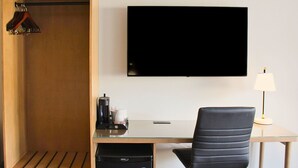 A hotel room with a wooden wardrobe, a desk with a chair, a flat-screen TV, a lamp, and a coffee maker.