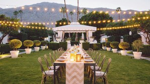 Un área de comedor al aire libre con una larga mesa dispuesta para una comida, rodeada de sillas y decorada con luces colgantes.