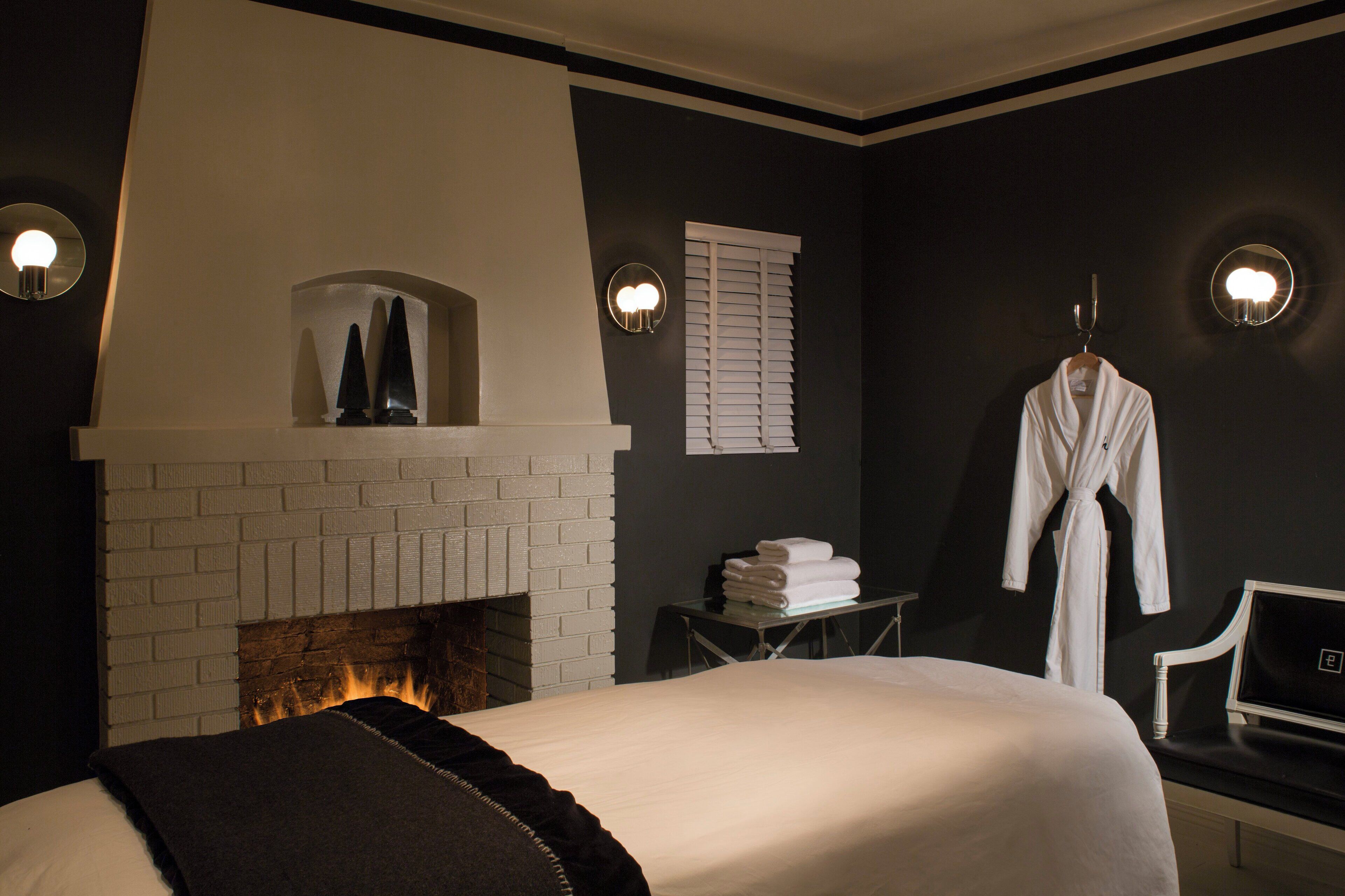 A hotel room with a white brick fireplace, a bed with a black throw, a robe hanging on a hook, and a desk with a laptop.