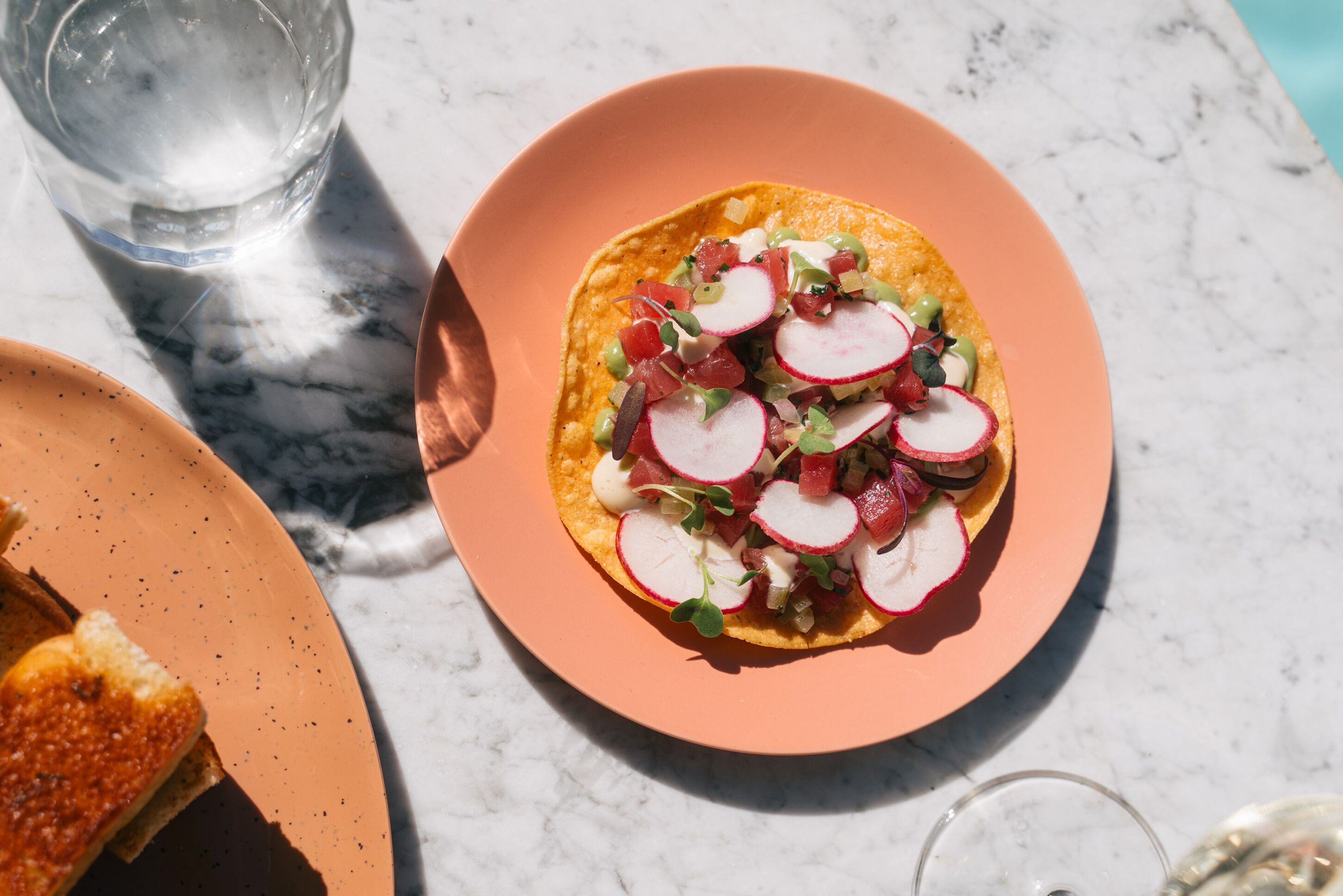 A plate with a tortilla topped with radishes and greens, a glass of water, and a slice of bread on a marble table.