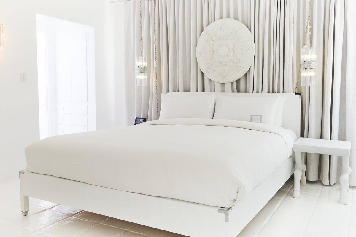 Une chambre à coucher blanche avec un lit, des rideaux et une table de chevet.