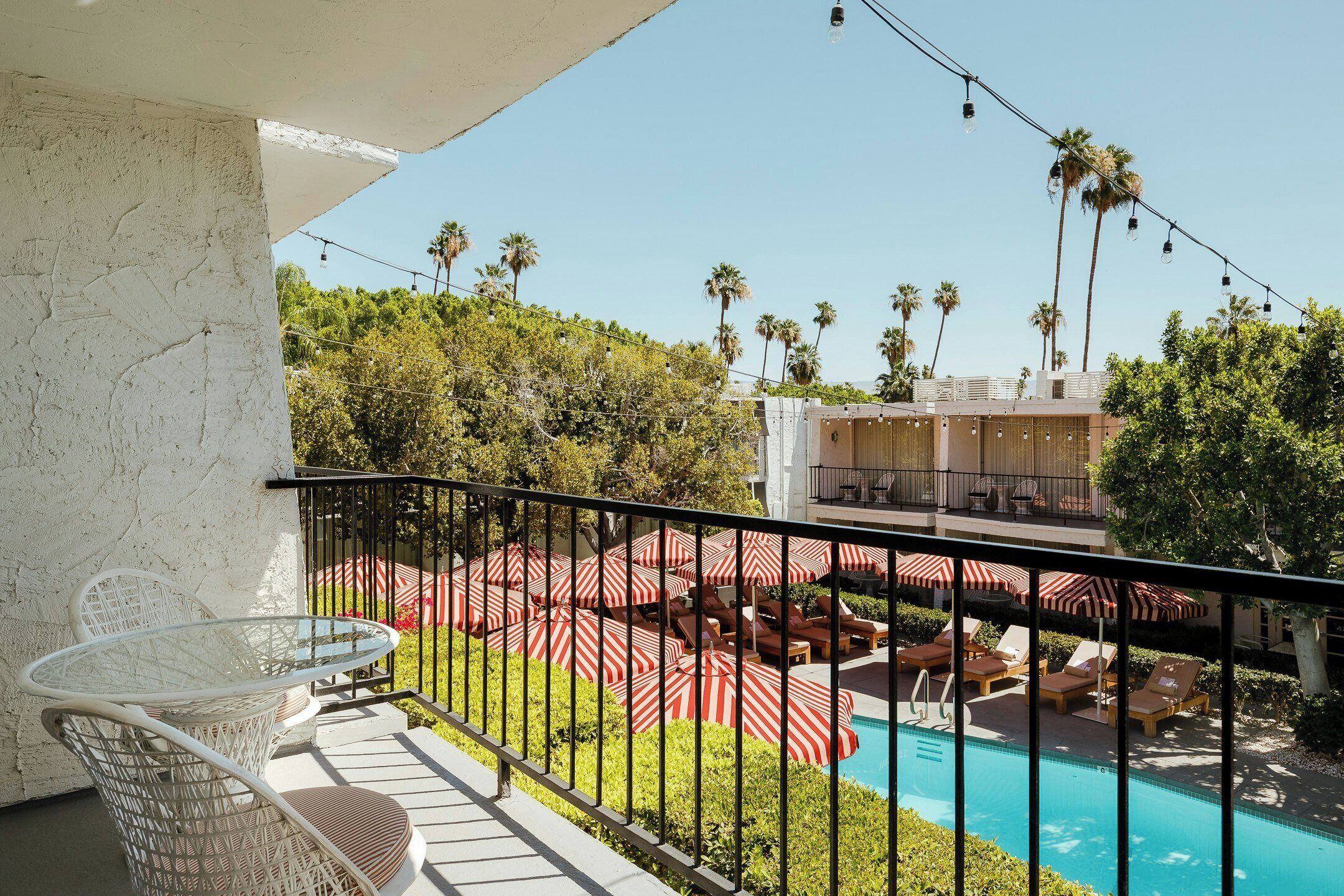 A balcony with a glass table and chairs, a pool area with sun loungers and umbrellas, and a view of palm trees and a building.