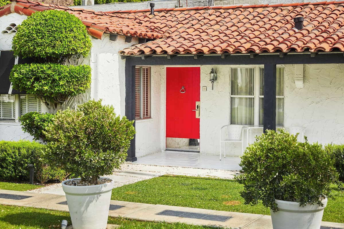 Un bâtiment avec une porte rouge, des murs blancs et un toit recouvert de tuiles. On aperçoit des plantes en pot au premier plan.