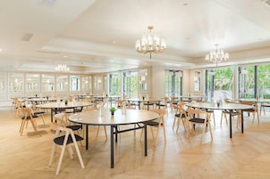 A spacious room with multiple round tables and chairs, large windows, and chandeliers.