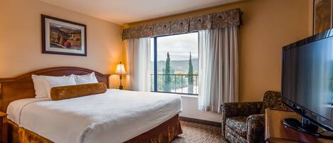 Habitación de hotel con una cama grande, televisión, una silla y ventana con vistas a árboles y colinas.
