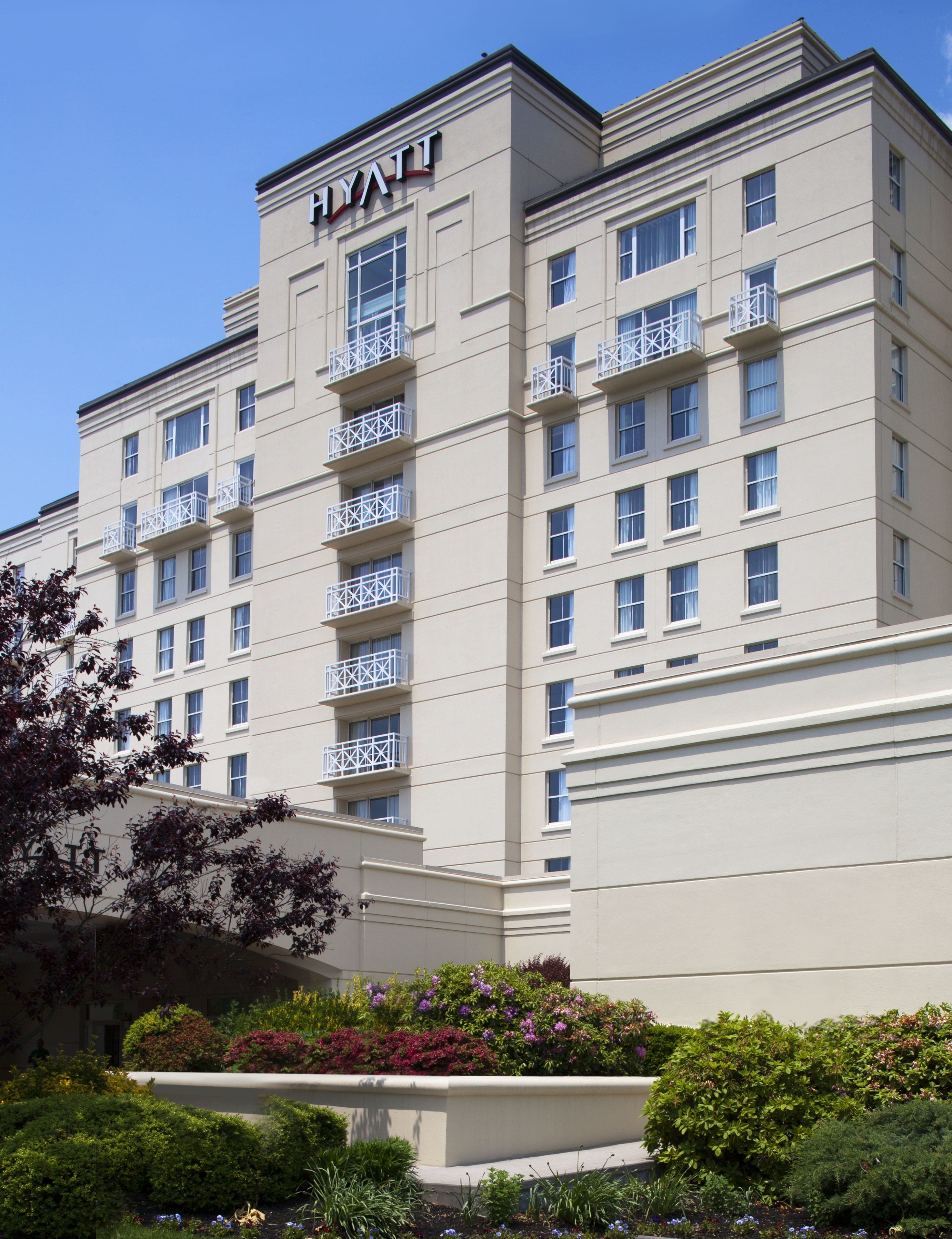 A multi-story hotel with a flat facade, balconies on each floor, and a landscaped garden area in front.