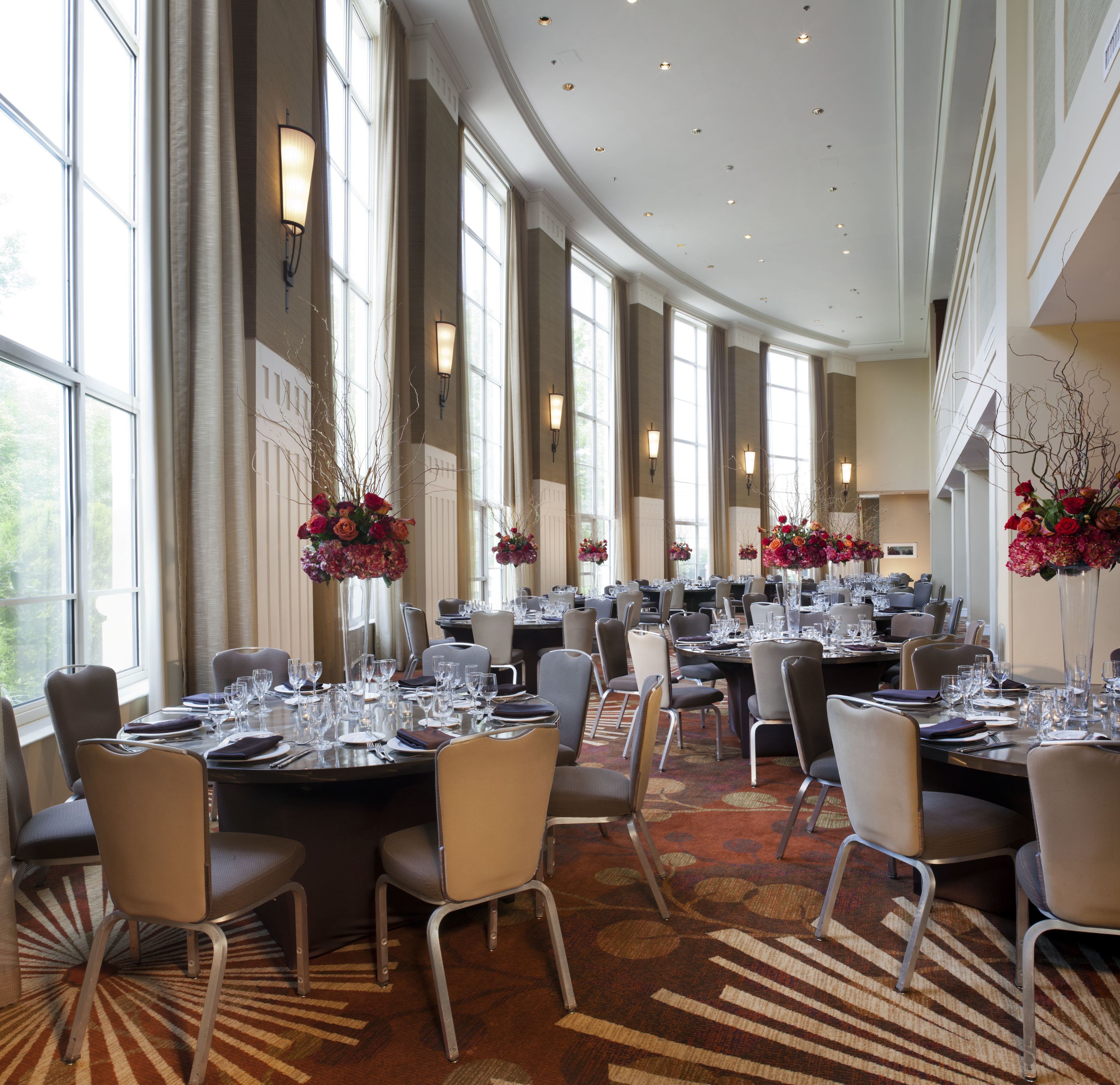 A large, elegantly furnished dining area with high ceilings and large windows, featuring multiple tables set for dining.