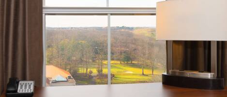 A hotel room with a desk, a lamp, a telephone, and a view of a golf course through a window.