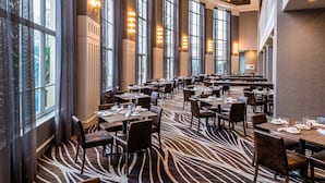 A spacious dining area with high ceilings, large windows, and a zebra-striped carpet. Tables are neatly arranged with chairs, and the room is well-lit with ceiling lights. - Hyatt Regency Long Island (Hauppauge)