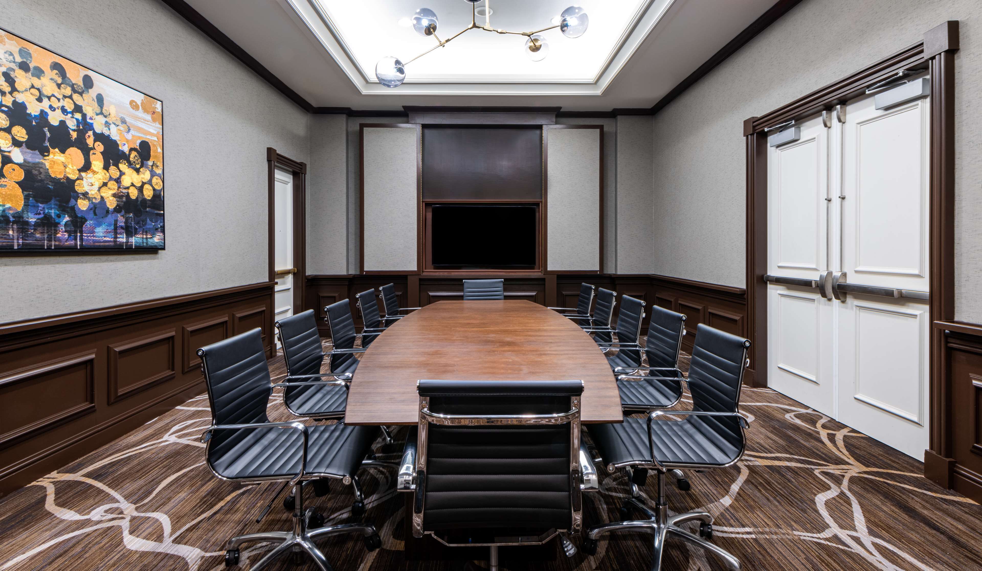 A conference room with a long table, black leather chairs, a large painting on the wall, and a ceiling light fixture.