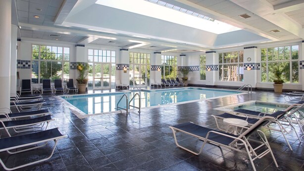 An indoor swimming pool with sunlit areas, lounge chairs, and potted plants. - Hyatt Regency Long Island (Hauppauge)