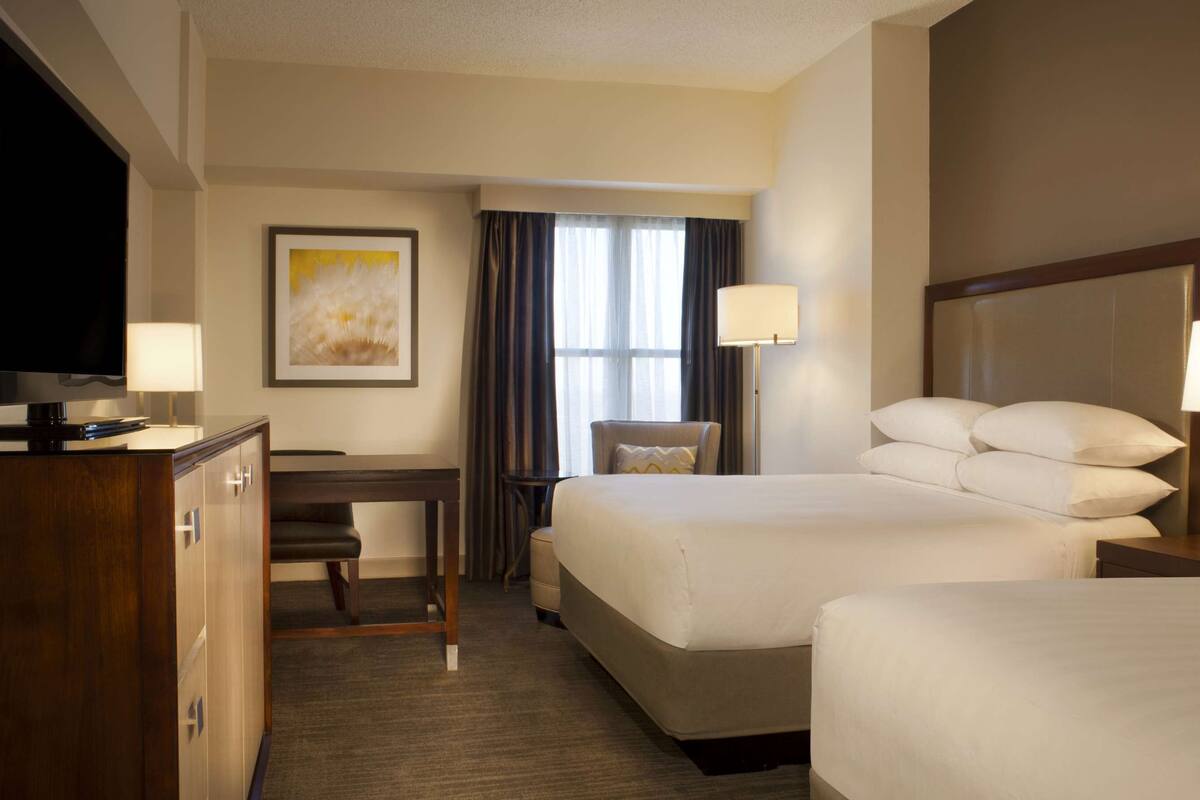 A hotel room with two beds, a desk, a chair, a television, and a painting on the wall.