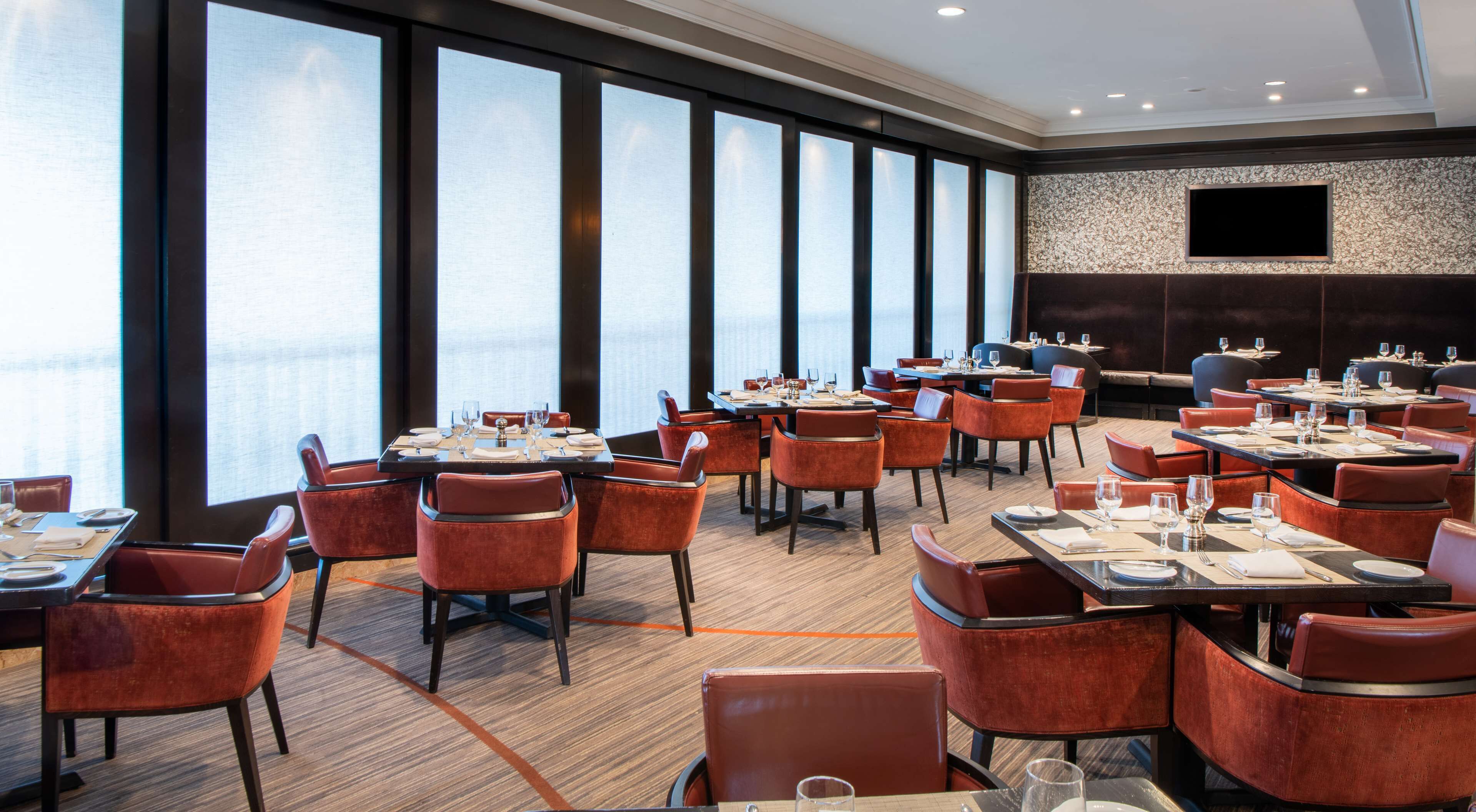 A modern dining area with multiple tables and chairs, large windows, and a stone wall with a flat-screen TV.