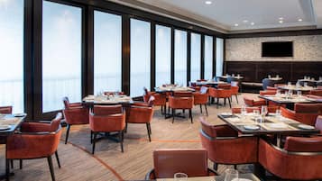 A modern dining area with multiple tables and chairs, large windows, and a stone wall with a flat-screen TV.