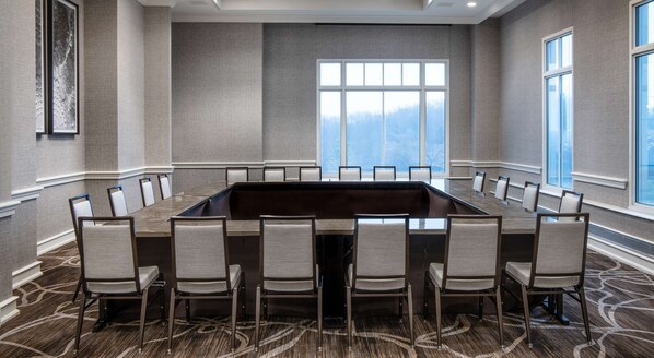 Un salón de conferencias con una amplia mesa rectangular rodeada de sillas, ventanas grandes con vistas a árboles y suelo alfombrado.