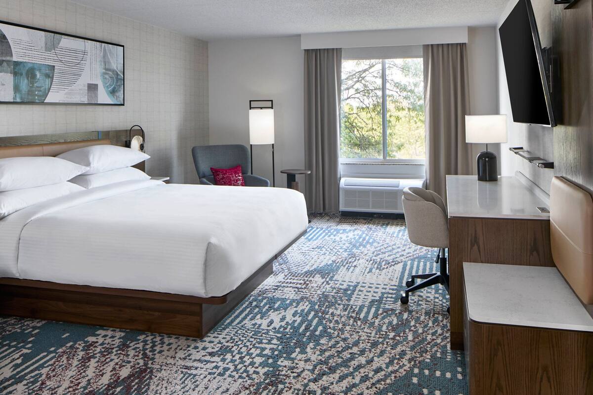 Ein Hotelzimmer mit einem großen Bett, einem Schreibtisch, einem Stuhl, einem Fernseher und einem Fenster mit Vorhängen.
