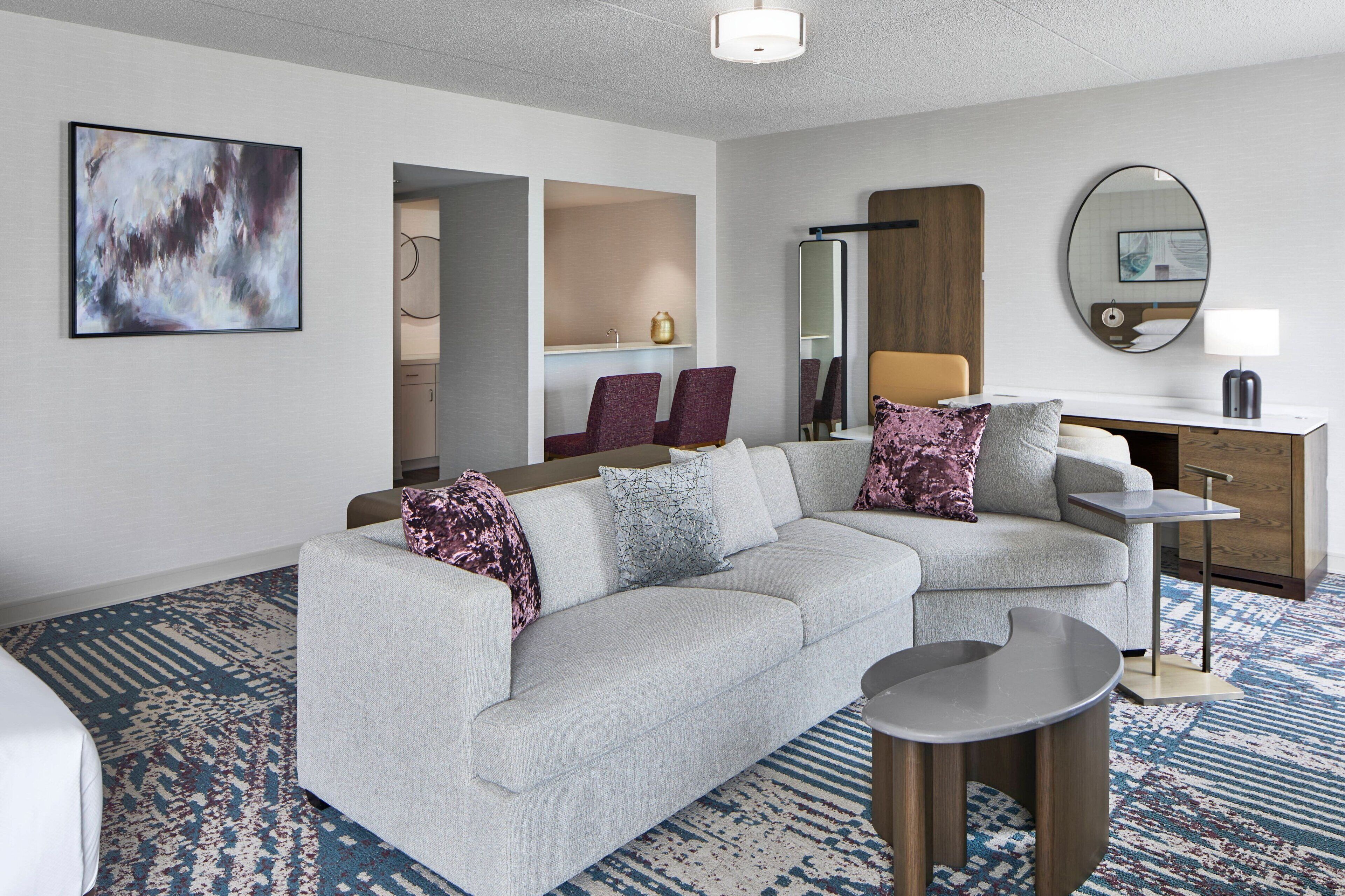 Habitación de hotel moderna con un sofá gris, una mesita redonda pequeña, un escritorio con lámpara, un cuadro abstracto grande y un espejo en la pared.