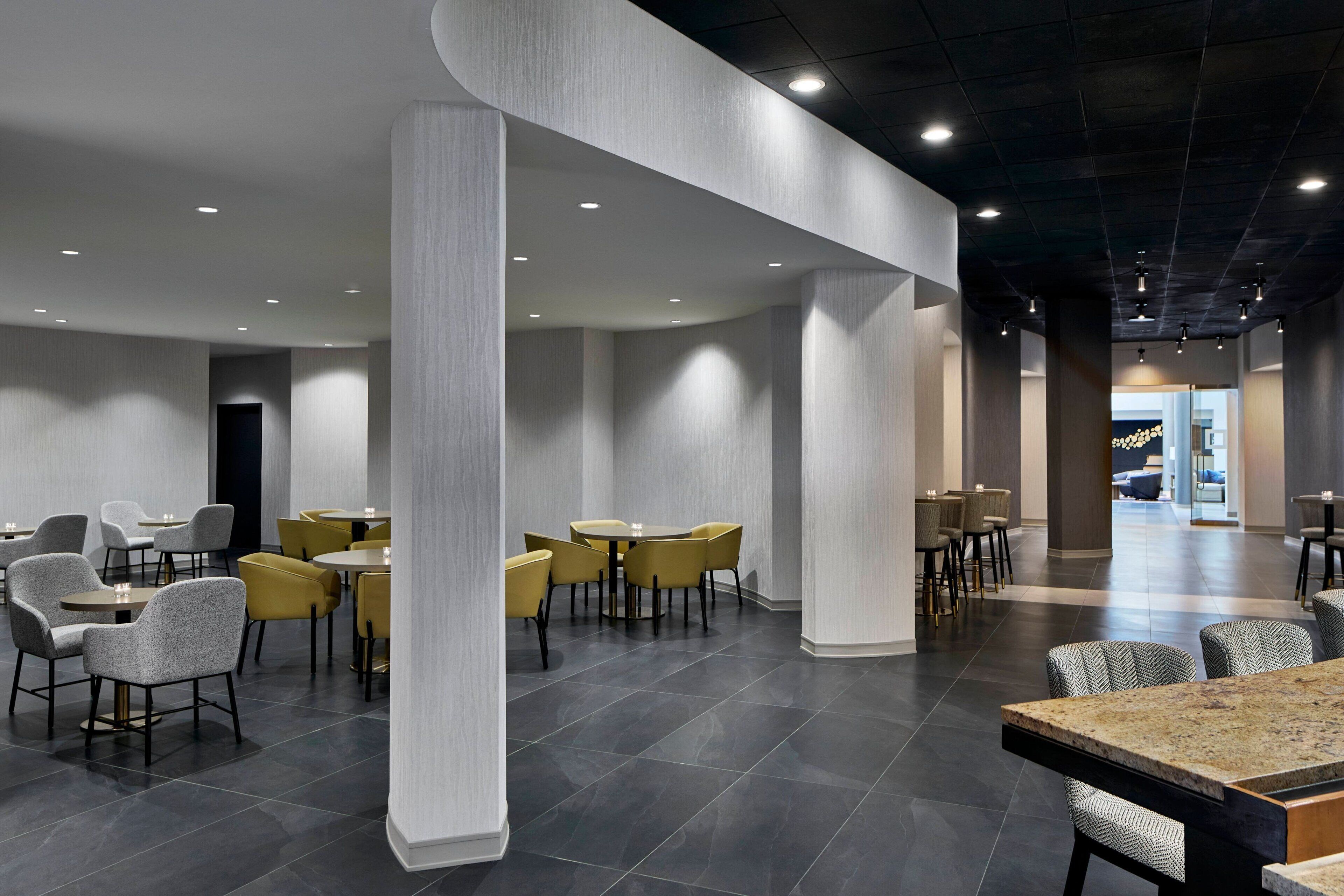 Un hall d’hôtel moderne avec un sol carrelé foncé, un espace salon central doté de fauteuils jaunes et gris, et une grande colonne.