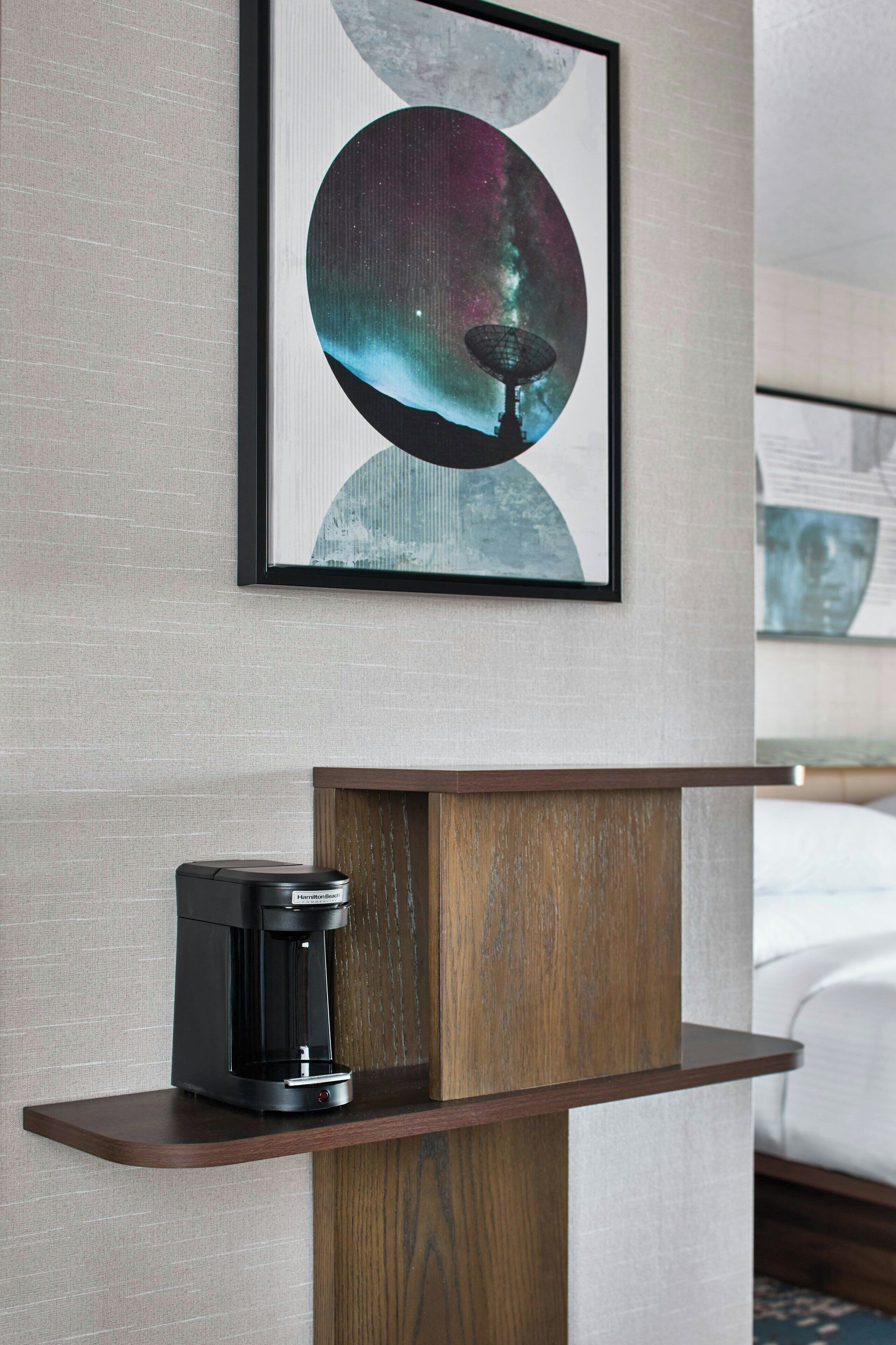 Ein gerahmtes Kunstwerk zeigt eine kosmische Szene mit einem Teleskop, einer Kaffeemaschine auf einer Holzablage und einem Bett im Hintergrund.