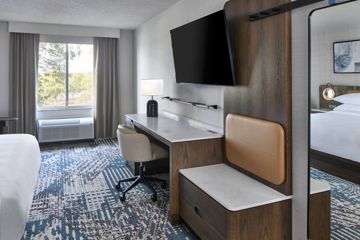 Ein Hotelzimmer mit Bett, Schreibtisch, Sessel, Fernseher und einem Fenster mit Vorhängen.