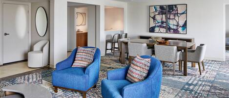 A modern living room with two blue armchairs, a dining area with a table and chairs, and a large abstract painting on the wall.