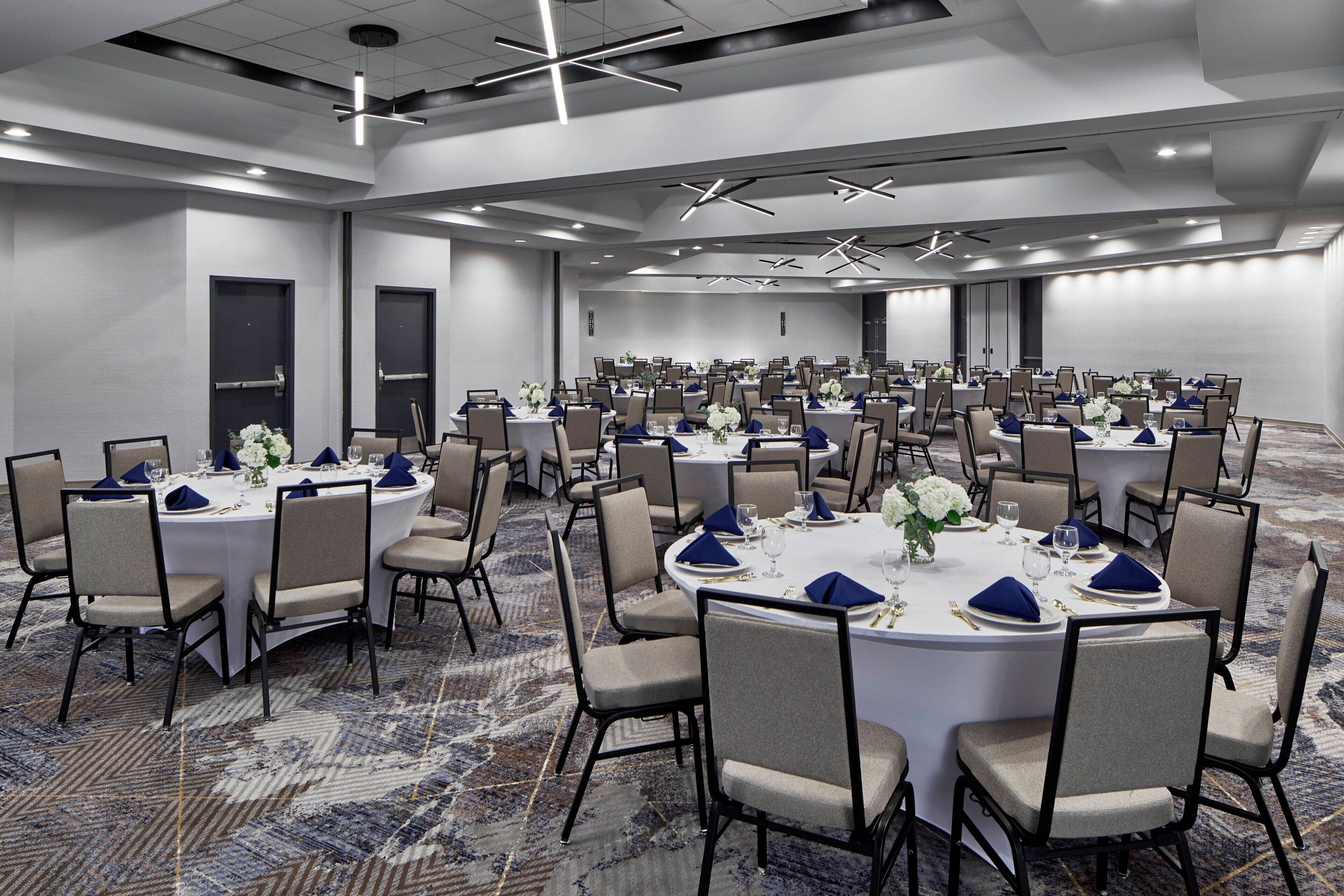 A large conference room with multiple round tables, each set with chairs, white tablecloths, and blue napkins.
