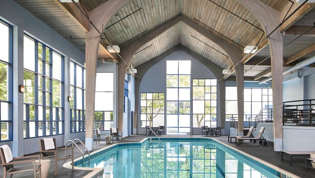 Una piscina interior con vistas despejadas al exterior gracias a amplios ventanales, un techo alto con vigas de madera y sillones de descanso a orillas de la piscina.