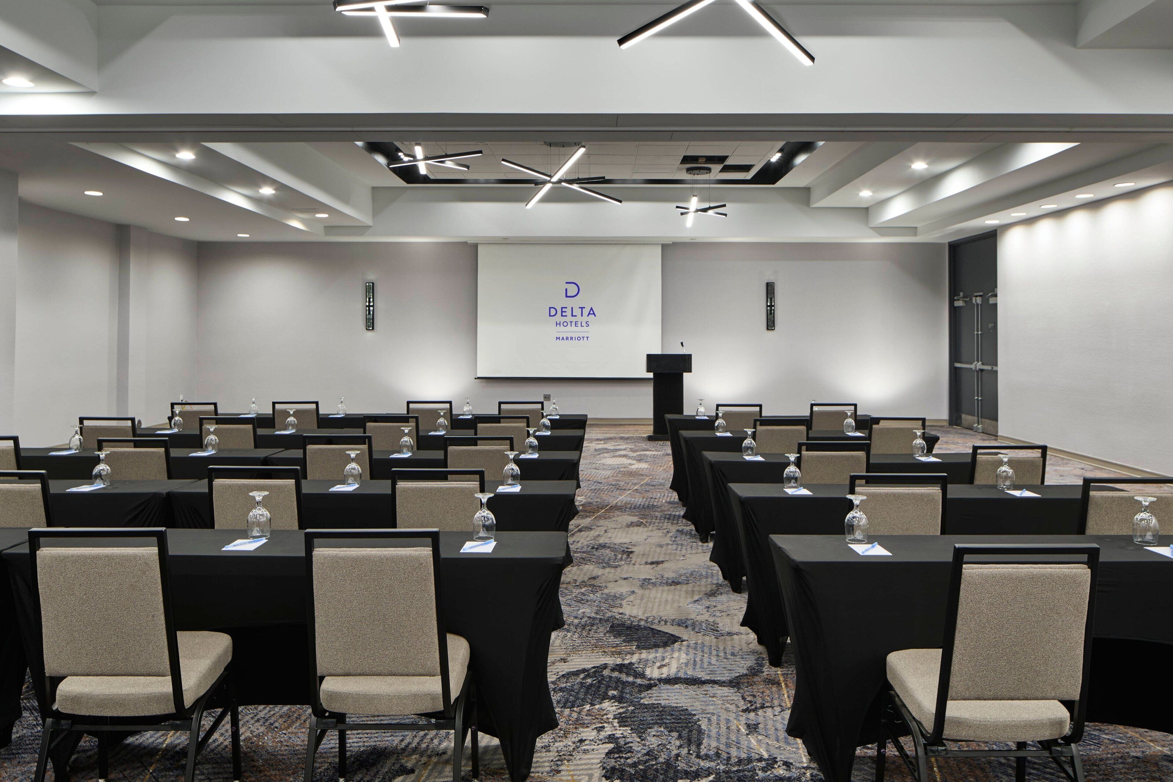 A conference room with rows of tables and chairs, a podium, and a screen displaying the Delta Hotels Marriott logo.