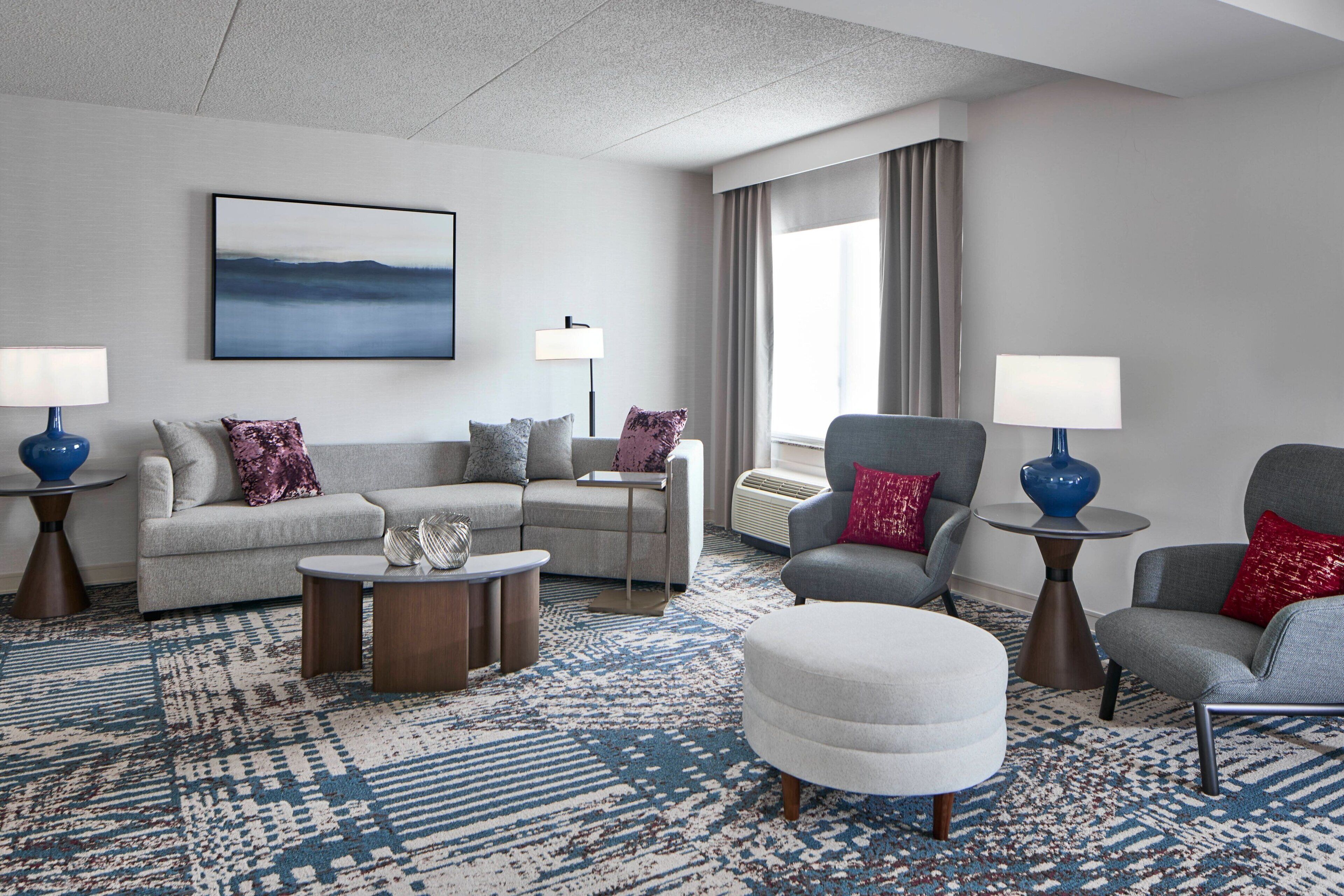 A modern hotel room with a grey sofa, armchairs, a round coffee table, and a large window with curtains.