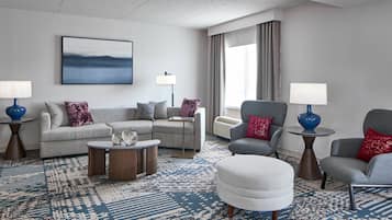 A modern hotel room with a grey sofa, armchairs, a round coffee table, and a large window with curtains.