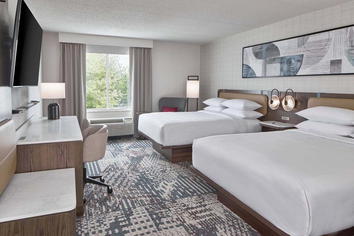 Ein Hotelzimmer mit zwei Betten, einem Schreibtisch, einem Sessel, einem Fernseher und einem großen Fenster mit Vorhängen.