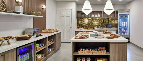 A modern kitchen area with a central island, shelves with snacks, a refrigerator, and a door.